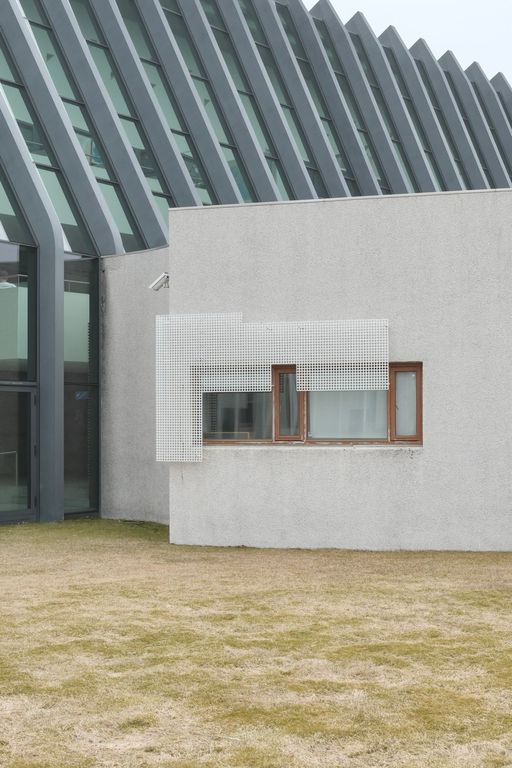 A photo of a building with a steel and glass main structure and a stucco stide building whose wooden-framed windows are partly covered by a perforated mesh panel. A security camera is mounted above the windows, with dry grass in the foreground.
